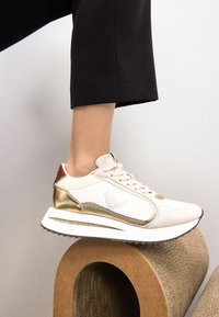 Zapatilla blanca con detalles en dorado y un talón rojo, con una parte superior de malla y cuero, suela blanca gruesa y detalles perforados en el costado.