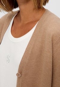 Ljusbrun cardigan med texturerad stickning, med en enda brun knapp. Bärs över en vit linne med ribbad halslinning och en broderad logotyp.