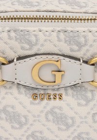 Borsa grigia chiara con tessuto testurizzato e un motivo sottile. Accessori in oro, incluso un prominente logo "G" e il marchio "GUESS". Chiusura a zip.