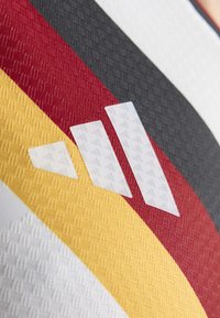 Witte trui met een gestructureerd patroon van strepen in rood, geel, grijs en zwart, met drie witte geometrische logo's die op het ontwerp zijn geplaatst.