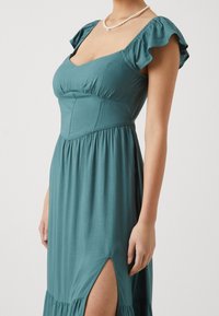 Tyrkysové maxi šaty se čtvercovým výstřihem, volánkovými rukávy a bočním rozparkem. Vyrobené z lehké látky s nařaseným pasem a volánkovým lemem.