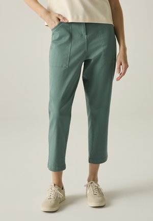 Mujer con pantalones verdes recortados con bolsillos delanteros y zapatillas beige, de pie con una mano en el bolsillo contra un fondo liso.