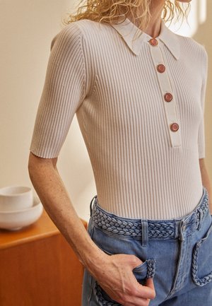 Kvinde iført en ribbet cremet kortærmet top med fire røde knapper og blå jeans med en flettet bælte og lomme detaljer.