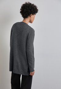 Cardigan grigio in maglia con vestibilità lunga e rilassata, polsini a coste e tasche laterali. La texture appare morbida e leggermente pelosa per offrire calore.