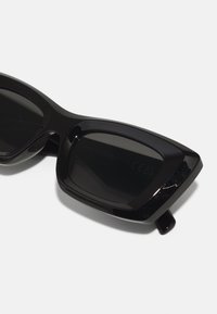 Lunettes de soleil noires avec une monture en plastique brillant, des verres rectangulaires et un design angulaire ; elles présentent une texture lisse et une construction légère.