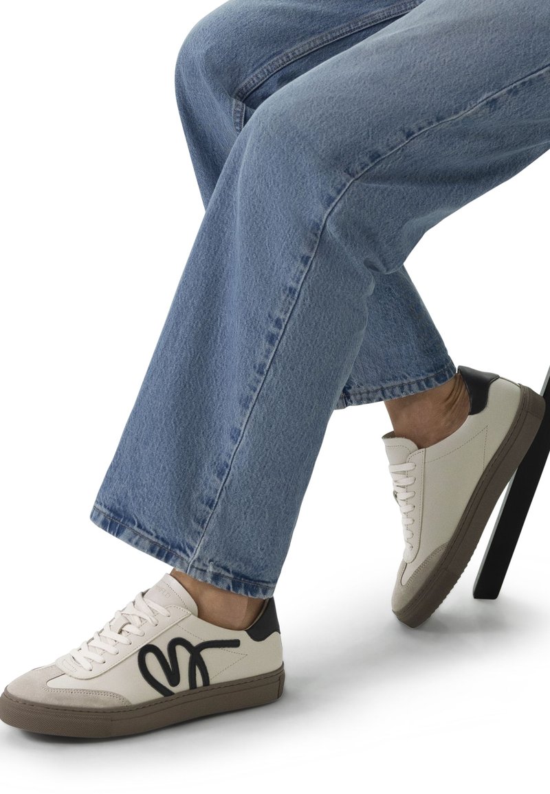 Baskets blanches avec accents noirs et un design graphique prononcé. Matériaux en daim et cuir, semelle en caoutchouc, portées avec un jean en denim bleu.