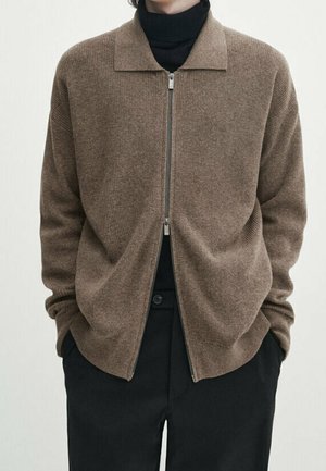 Cardigan marron à fermeture éclair porté par-dessus un col roulé noir, mains dans les poches d'un pantalon noir, visible du cou aux hanches.