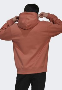 Man som justerar huvan på en rostfärgad hoodie, sedd bakifrån, bärande svarta byxor mot en ljus, enfärgad bakgrund.