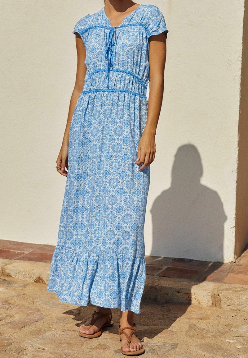 Femme portant une robe bleue à motifs jusqu'aux chevilles, manches courtes et sandales, debout à l'extérieur sur une surface en pierre près d'un mur blanc.