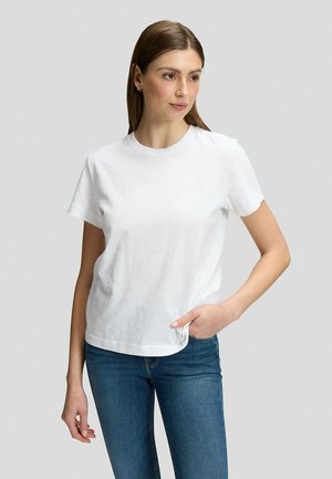 Frau mit glattem braunem Haar trägt ein schlichtes weißes T-Shirt mit kurzen Ärmeln und blaue Jeans, steht mit einer Hand in der Tasche.