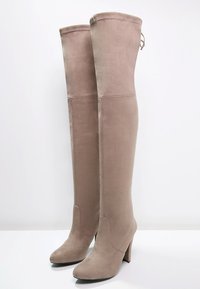 Bottes taupe en daim, au-dessus du genou, avec de hauts talons blocs et des orteils ronds, se tenant debout sur une surface blanche contre un fond uni.