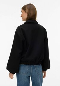 Veste noire surdimensionnée de style bomber avec un col, une taille élastique et des manches larges ; le modèle la porte avec un jean bleu.