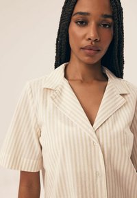 Koszula na krótki rękaw, z kołnierzykiem w jasnobeżowym kolorze, w pionowe paski. Posiada zapięcie na guziki i miękką, teksturowaną tkaninę.
