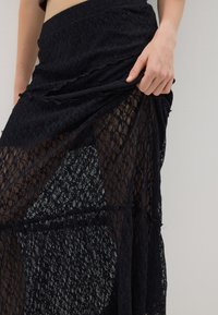 Schwarzer Spitzenrock mit durchsichtigen Einsätzen, strukturiertem Stoff, geschichteter Gestaltung und gerafftem Bund, der filigrane Muster und eine lange Form präsentiert.