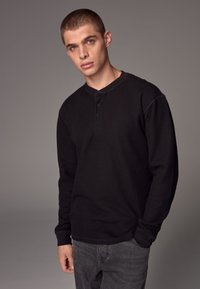 Czarna longsleeve koszula henley z prążkowanej tkaniny, z trzema guzikami przy dekolcie, połączona z ciemnoszarymi dżinsami, na neutralnym tle.
