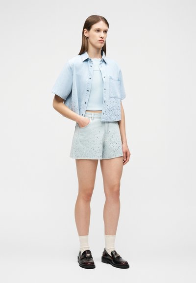 Γυναίκα όρθια με τα χέρια στις τσέπες, φορώντας ανοιχτό μπλε κοντομάνικο πουκάμισο, ταιριαστά βερμουδάκια με στίγματα, λευκές κάλτσες και μαύρα μοκασίνια.
