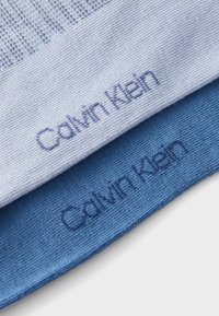 Dos muestras de tela en azul claro y azul oscuro, cada una con "Calvin Klein" bordado en colores a juego. Las texturas muestran patrones acanalados.