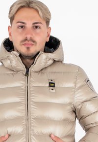 Man met kort lichtbruin haar, gekleed in een beige pufferjack met capuchon, zwarte ritssluiting en een 'Blauer U.S.A.' patch op de borst en schouder.