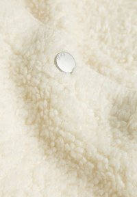 Tissu en polaire blanc avec une surface texturée et duveteuse. Présente une petite étiquette en métal argenté embossée avec "IKKS."