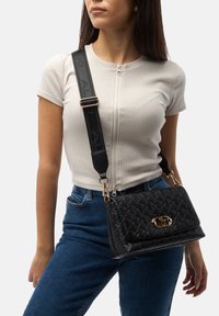 Borsa a tracolla nera trapuntata con accessori dorati, tracolla regolabile e finitura testurizzata. Indossata a tracolla, abbinata a un top beige aderente e jeans.