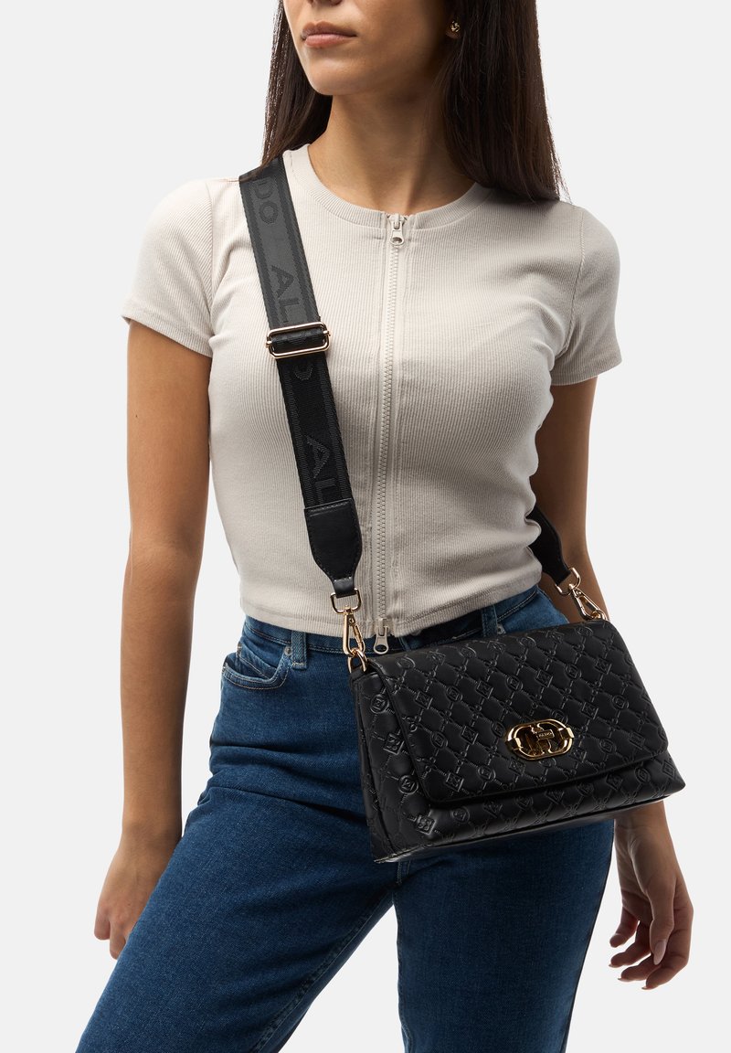 Borsa a tracolla nera trapuntata con accessori dorati, tracolla regolabile e finitura testurizzata. Indossata a tracolla, abbinata a un top beige aderente e jeans.