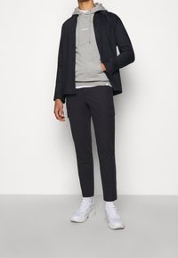 Conjunto masculino que incluye una chaqueta oscura, sudadera gris con cordones, pantalones negros ajustados y zapatillas blancas. Los materiales son texturizados y casuales.