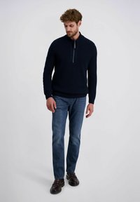 Marineblauwe gebreide trui met hoge kraag en kwartrits, gecombineerd met lichtblauwe rechte jeans en donkerbruine schoenen.