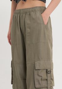 Cargo-Hosen aus olivgrünem Stoff, mit einem elastischen Bund, seitlichen Taschen und Cargo-Details an den Oberschenkeln mit genähten Akzenten.