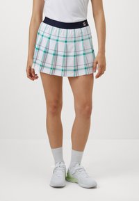Gonna bianca a scacchi con accenti verdi e blu navy, caratterizzata da un design plissettato e una vita solida blu navy. Indossata con sneakers bianche e calzini grigi.