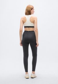 Αθλητικό σουτιέν σε κρεμ απόχρωση με μαύρη λωρίδα "Tommy Jeans"· συνδυασμένο με εφαρμοστά σκουρόγρια κολάν. Το μοντέλο φοράει ανοιχτόχρωμα παπούτσια. Μινιμαλιστικός σχεδιασμός.