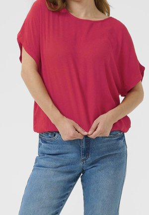 Femme portant un haut rouge ample à manches courtes avec un jean bleu, tenant l'ourlet du haut avec les deux mains à la taille.