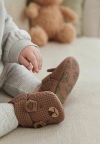 Papuci pentru bebeluși maro cu textură, având accente în formă de față de urs și curele cu arici, cu tălpi verzi cu un model. Îmbrăcăminte de culoare gri deschis cu dungi.