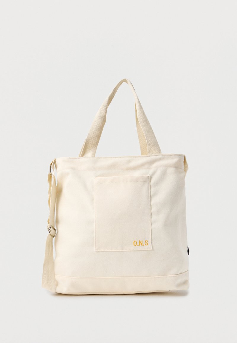 Sac fourre-tout en toile crème avec poche avant, deux poignées, bandoulière réglable et petit texte jaune "O.N.S" en bas à droite de la poche.