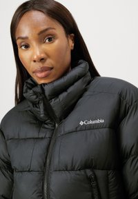 Geacă puffer neagră cu guler înalt, model matlasat orizontal, fermoar frontal și logo Columbia pe piept. Material moale și izolat.