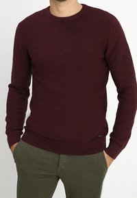 Homme portant un pull texturé à manches longues rouge foncé et un pantalon vert olive, les mains dans les poches, devant un fond uni.