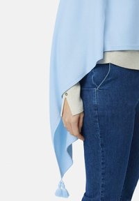 Dłoń oparta wzdłuż boku, ubrana w beżowy, prążkowany sweter z długim rękawem oraz jasnoniebieski szal z frędzlami, połączone z ciemnoniebieskimi dżinsami.