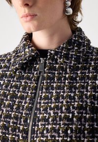 Close-up van een persoon die een gestructureerd zwart, paars en groen jack met rits draagt en grote zilveren oorbellen met dubbele bol.