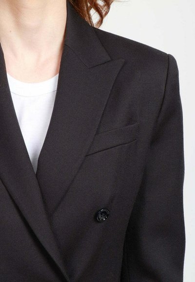 Blazer nero doppiopetto con rever a lancia, tessuto texturizzato, una tasca sul petto e dettaglio del bottone nero. Indossato sopra una maglietta bianca.