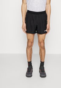 Hombre vistiendo pantalones cortos deportivos negros, calcetas negras hasta el tobillo, zapatillas gris oscuro con acentos naranjas y una camiseta sin mangas blanca de malla.