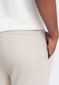 Pantalones beige texturizados con un bolsillo trasero plano y un bolsillo lateral. La tela parece transpirable, con un acabado suave.