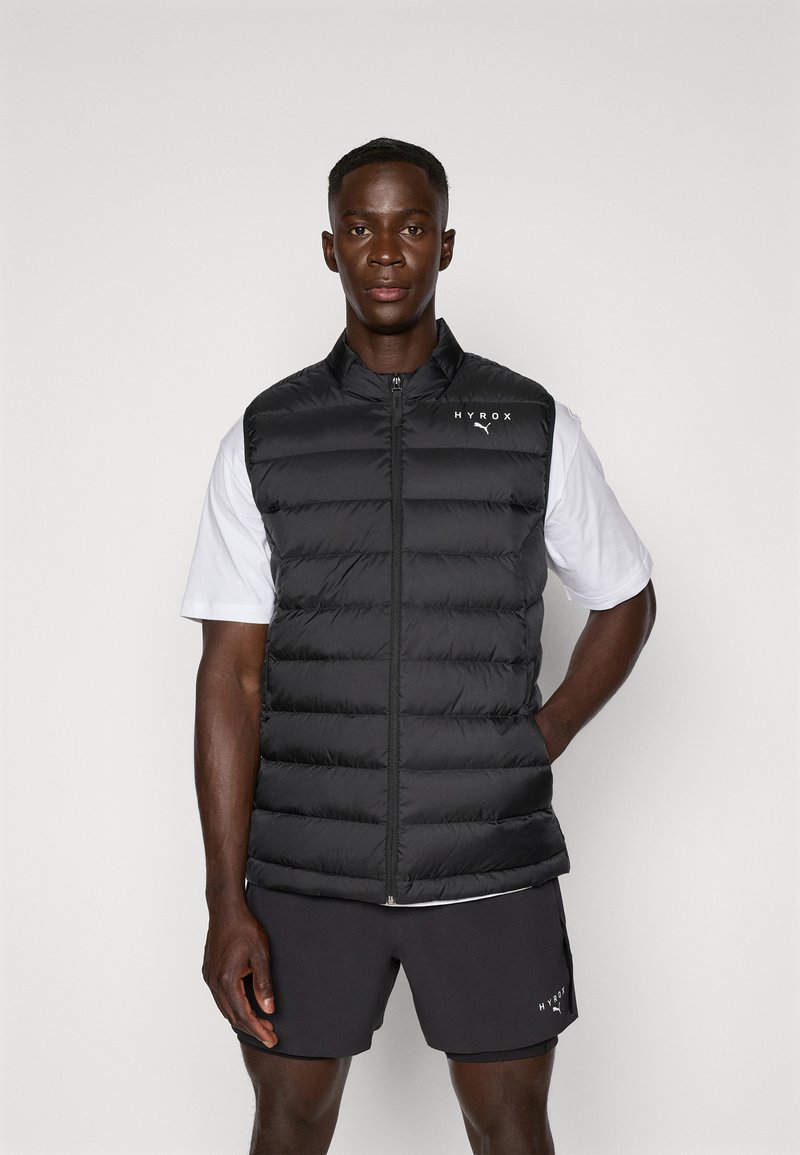 Gilet matelassé noir avec un col haut, un design en quilt et une fermeture éclair à l'avant. Présente le logo blanc "HYROX" sur la zone gauche de la poitrine.