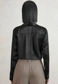 Person mit glattem schwarzen Haar, der ein schwarzes Satin-Hemd mit langen Ärmeln trägt, das in hochgeschnallte beige Hosen gesteckt ist, von hinten gezeigt.