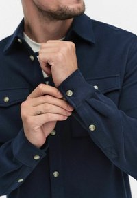 Marineblå skjorta med en texturerad tyg, som har knäppt manschetter, bröstfickor och kontrasterande ljusa knappar. Händer som justerar manschetten.