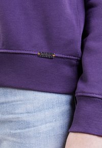 Fialová mikina s ribovaným spodním lemem a manžetami. Obsahuje štítek přišitý na spodní části, doplňující světle modré džíny v pozadí.