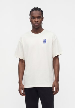 Junger Mann mit geflochtenem Haar, der ein weißes übergroßes T-Shirt mit kleinem blauem Motiv auf der linken Brust und schwarze Hose trägt, steht.