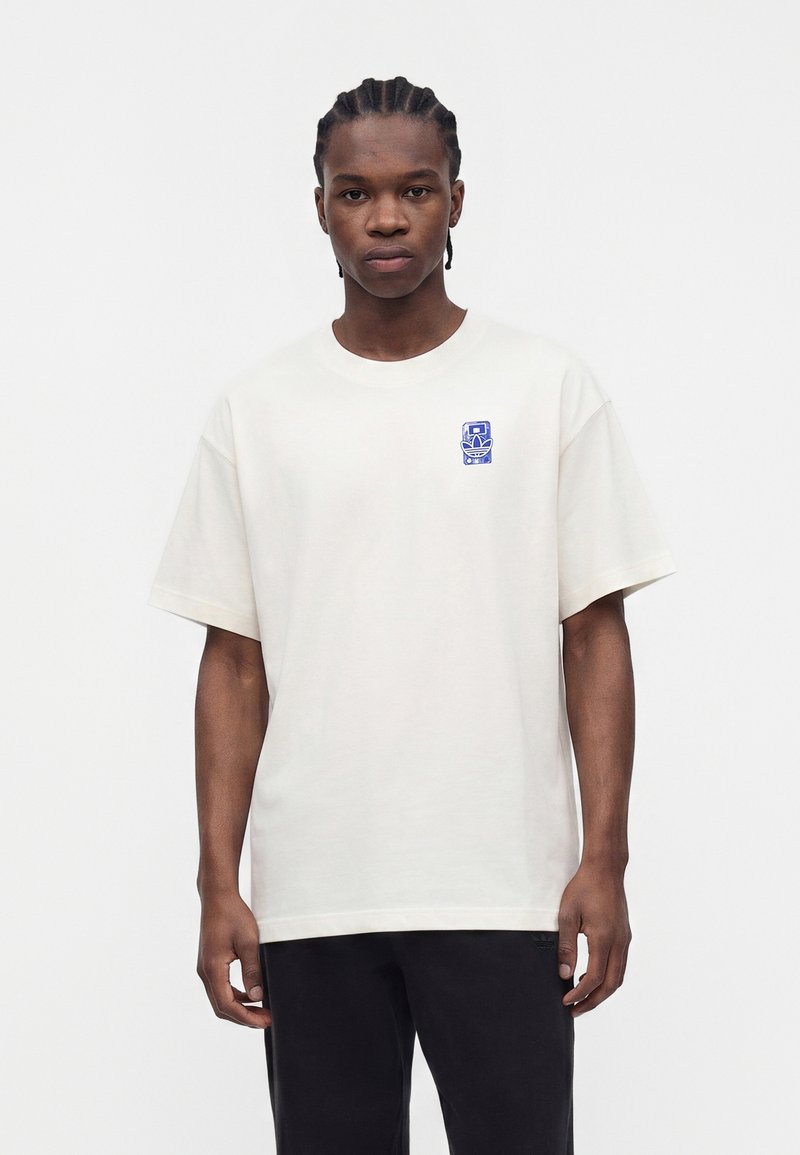 Jeune homme aux cheveux tressés portant un t-shirt blanc oversize avec un petit motif bleu sur la poitrine gauche et un pantalon noir, debout.
