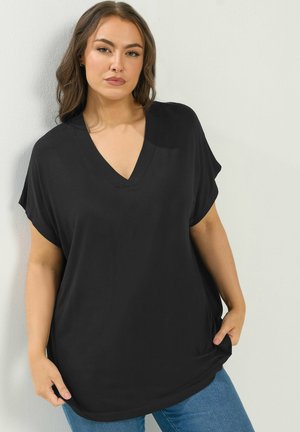 Frau mit braunen Haaren, die ein lockeres schwarzes V-Ausschnitt-Oberteil und blaue Jeans trägt, steht vor einer hellgrauen Wand.