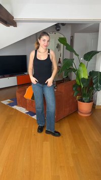 Γυναίκα με μαύρο τοπ χωρίς μανίκια και μπλε τζιν στέκεται χαμογελώντας σε ένα σαλόνι με καφέ καναπέ, μεγάλο γλαστρόφυτο και ξύλινο πάτωμα.