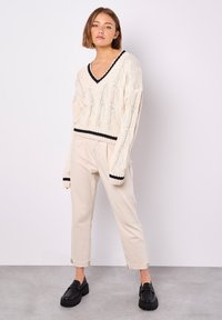 Hellbeige gestrickter Pullover mit V-Ausschnitt, schwarzer Einfassung und lockerer Passform. Kombiniert mit hellbeigen Hosen und schwarzen Plateauschuhen.