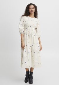 Robe midi crème avec des manches bouffantes, un corsage smocké et des broderies florales multicolores. Portée avec des bottines noires.